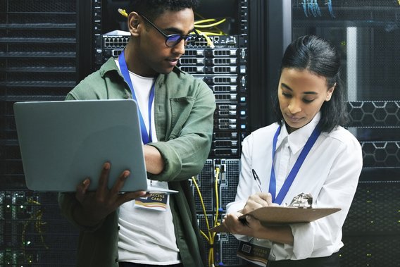 Zwei IT-Fachkräfte prüfen mit Laptop und Klemmbrett Server in einem Rechenzentrum.