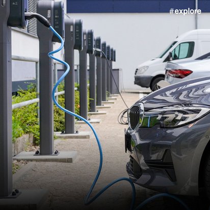 Ein graues Elektrofahrzeug lädt an einer öffentlichen Ladestation mit blauem Kabel in einem Wohngebiet. Im Hintergrund sind weitere Fahrzeuge und Wohnhäuser mit roten Backsteinfassaden zu sehen.