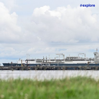 Ein großes Schiff oder eine Offshore-Plattform in der Ferne, fotografiert über eine Grasfläche mit einem bewölkten Himmel im Hintergrund.