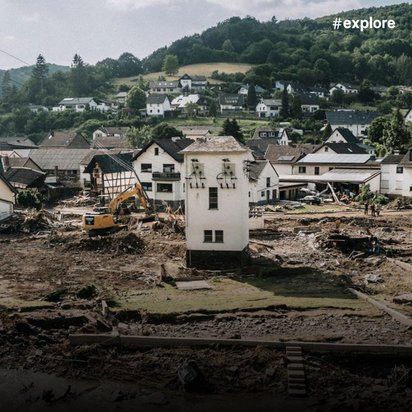 Dorflandschaft mit schwerem Hochwasserschaden: zerstörte Häuser und Schlammmassen im Vordergrund, intakte Gebäude im Hintergrund auf Anhöhen.