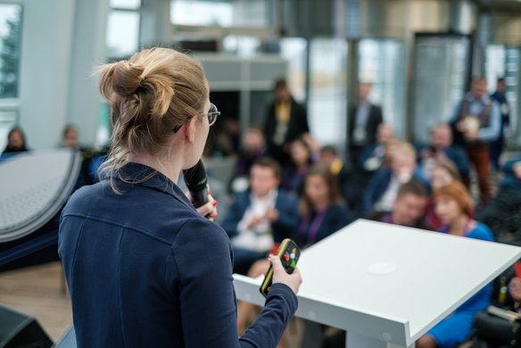 Frau mit Mikrofon spricht vor Publikum auf einer Konferenz oder Tagung, Blick von hinten auf Bühne und Zuhörer
