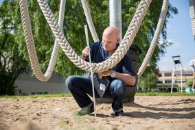 Ein Mann in dunkler Arbeitskleidung kniet neben dicken Seilen und misst mit einem Maßstab den Abstand zwischen Spielgeräten. Im Hintergrund sind große runde Spielelemente und grüne Bäume zu sehen.
