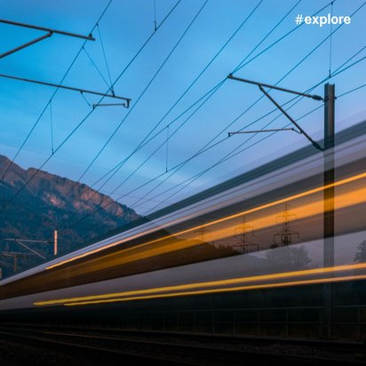 Langzeitbelichtung eines vorbeifahrenden Zuges in der Dämmerung, wodurch gelb-orange Lichtstreifen entstehen. Die Aufnahme zeigt Oberleitungen vor einer bergigen Landschaft mit violett-blauem Himmel.