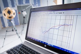 Laptop-Bildschirm zeigt grafische Messdaten mit roten und blauen Linien; im Hintergrund ist ein kugelförmiges Messinstrument auf einem Stativ zu sehen.