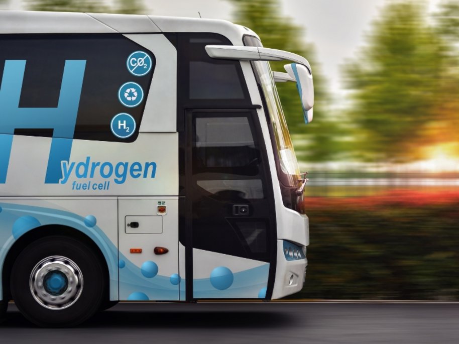 Weißer Wasserstoff-Brennstoffzellen-Bus mit blauer "Hydrogen fuel cell" Beschriftung und Umweltsymbolen fährt bei Bewegungsunschärfe durch grüne Landschaft.
