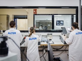 Drei Wissenschaftler in weißen ALTER-Laborkitteln arbeiten an modernen Geräten in einem sterilen Forschungslabor.