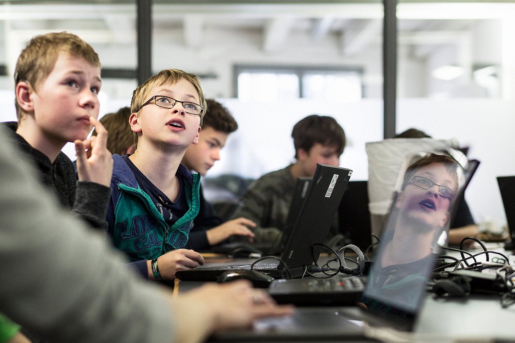 Eine Gruppe Jungen an Computern in einem hellen Klassenraum, wobei ein Junge mit Brille aufmerksam nach oben schaut.