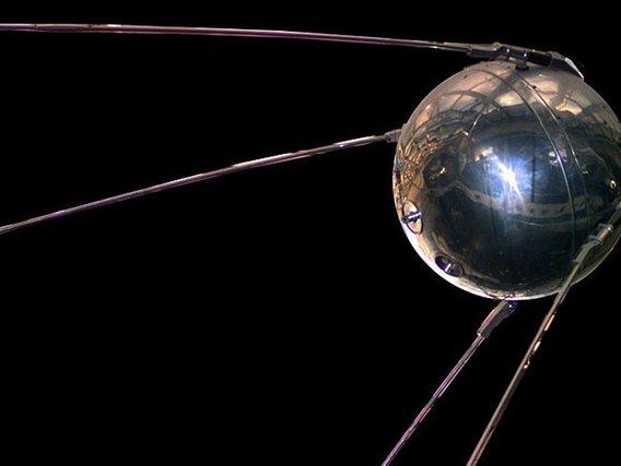 Erster künstlicher Satellit Sputnik als Metallkugel mit vier abstehenden Antennen vor schwarzem Hintergrund.