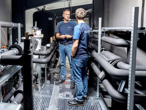 Two TÜV employees stand in a narrow corridor surrounded by pipes.
