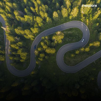 Eine geschwungene Straße schlängelt sich durch einen dichten, grünen Wald. Aus der Vogelperspektive sind mehrere scharfe Kurven zu sehen, mit wenigen Autos, die sich durch die Landschaft bewegen. Sonnenlicht fällt auf die Baumwipfel und erzeugt ein Spiel aus Licht und Schatten.
