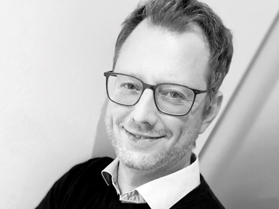 Ein schwarz-weißes Porträt von Dr. Jan Hammer mit schwarzer Brille, Hemd und Pullover