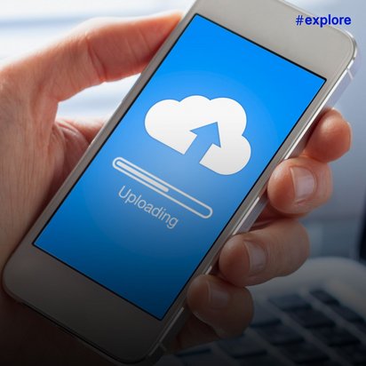 Eine Hand hält ein Smartphone mit blauem Bildschirm, der ein Cloud-Symbol mit Pfeil und "Uploading" Text sowie einen Ladebalken zeigt. Im Hintergrund ist unscharf ein Laptop auf einem Schreibtisch zu erkennen.