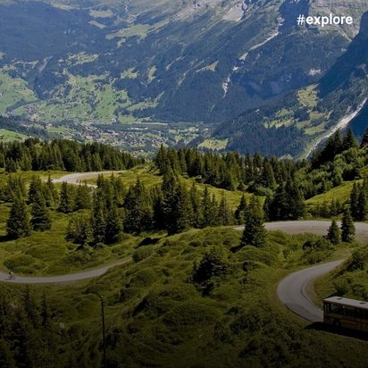 Spektakuläre Alpenlandschaft mit einem gelben Bus, der eine kurvige Bergstraße durch grüne Almwiesen hinunterfährt, umgeben von dramatischen Berggipfeln und einem Tal mit Ortschaft im Hintergrund.