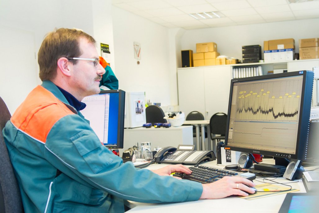 Eine Person sitzt in einem Büro an einem Schreibtisch und arbeitet an einem Computer. Auf dem Bildschirm ist ein Diagramm mit Kurven und Datenpunkten zu sehen. Im Hintergrund stehen mehrere weiße Schränke, auf denen Kartons und Ordner gestapelt sind. Auf dem Schreibtisch befinden sich eine Tastatur, eine Maus, ein Telefon und einige Büromaterialien. Die Person trägt eine Arbeitsjacke in Grün und Orange.