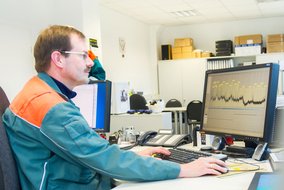 Eine Person sitzt in einem Büro an einem Schreibtisch und arbeitet an einem Computer. Auf dem Bildschirm ist ein Diagramm mit Kurven und Datenpunkten zu sehen. Im Hintergrund stehen mehrere weiße Schränke, auf denen Kartons und Ordner gestapelt sind. Auf dem Schreibtisch befinden sich eine Tastatur, eine Maus, ein Telefon und einige Büromaterialien. Die Person trägt eine Arbeitsjacke in Grün und Orange.