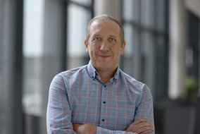 Eine Person steht mit verschränkten Armen in einem modernen Bürogebäude. Sie trägt ein kariertes Hemd, und der Hintergrund zeigt große Fenster, die viel Licht hereinlassen.