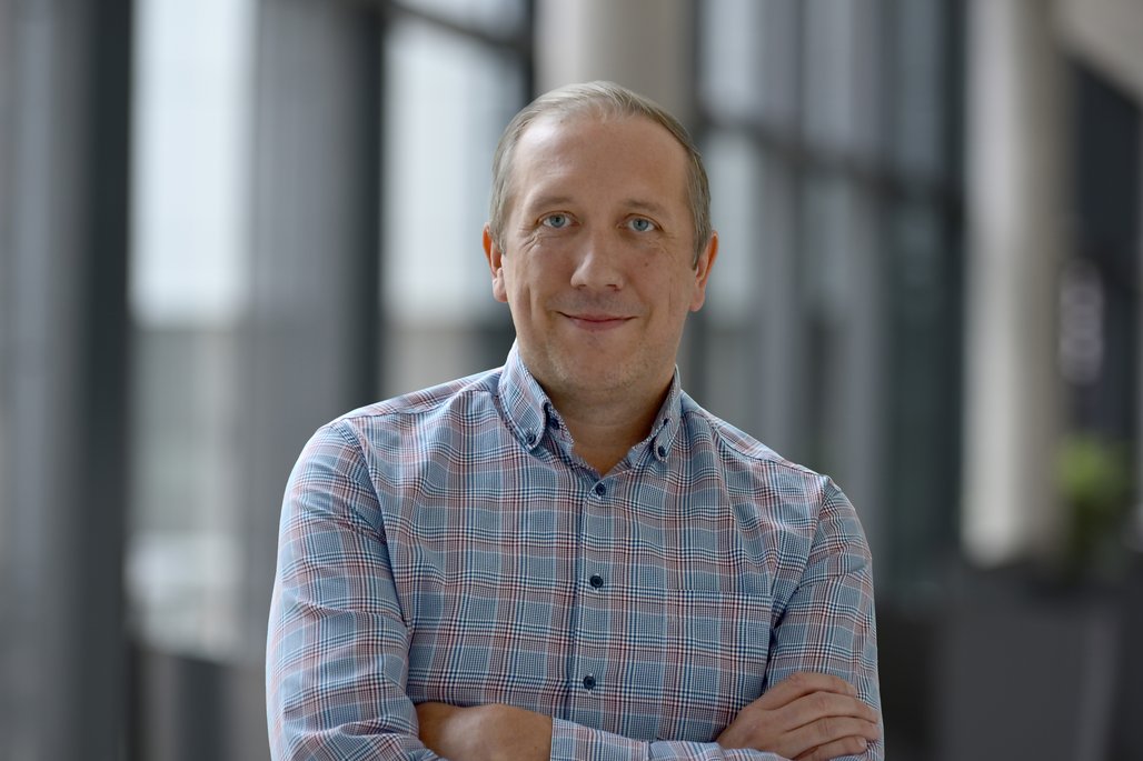 Eine Person steht mit verschränkten Armen in einem modernen Bürogebäude. Sie trägt ein kariertes Hemd, und der Hintergrund zeigt große Fenster, die viel Licht hereinlassen.
