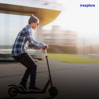 Dynamische Bewegungsaufnahme eines jungen Mannes in kariertem Hemd und Mütze, der mit einem E-Scooter an einem modernen Bahnhof vorbeifährt. Die Bewegungsunschärfe und das warme Sonnenlicht erzeugen ein Gefühl von Geschwindigkeit und urbaner Mobilität.