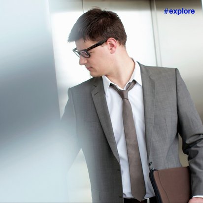 Ein junger Mann in einem grauen Anzug mit weißem Hemd und grauer Krawatte trägt schwarze Brille und wartet auf den Aufzug.