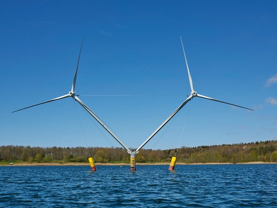 Two offshore wind turbines positioned in calm blue water, each mounted on yellow cylindrical foundations. The turbines feature white towers and three-blade rotors against a clear sky with forested shoreline in the distance.