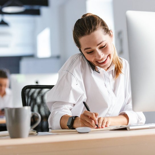 Lächelnde Frau telefoniert am Schreibtisch und macht Notizen am Computer