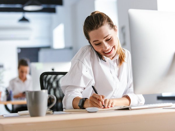 Lächelnde Frau telefoniert am Schreibtisch und macht Notizen am Computer