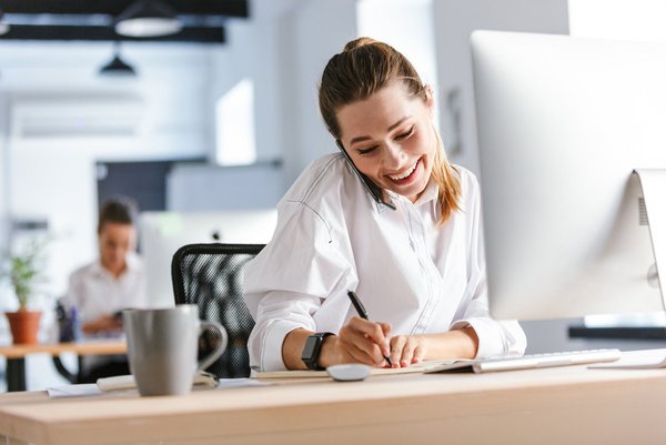 Lächelnde Frau telefoniert am Schreibtisch und macht Notizen am Computer