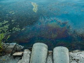 Blick von oben auf verschmutztes Gewässer mit Öl- oder Algenschichten auf der Oberfläche und zwei grauen Betonrohren am steinigen Ufer.