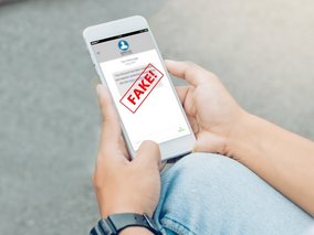 Ein weißes Smartphone in den Händen einer Person zeigt einen Bildschirm mit einem großen roten "FAKE!"-Stempel über einer Nachricht.