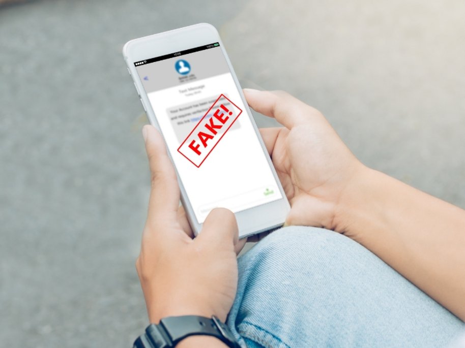 Ein weißes Smartphone in den Händen einer Person zeigt einen Bildschirm mit einem großen roten "FAKE!"-Stempel über einer Nachricht.