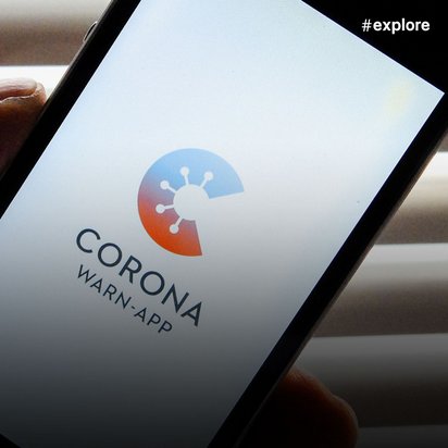 Eine Hand hält ein Smartphone mit der Corona-Warn-App auf dem Display, die ein stilisiertes Virus-Symbol in blau-rot zeigt. Die App wird vor einem hellen Jalousien-Hintergrund präsentiert und dient der digitalen Kontaktverfolgung während der Corona-Pandemie.