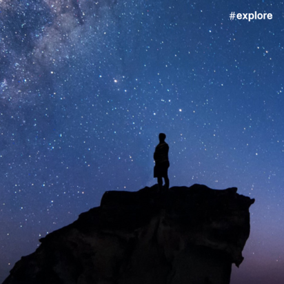 Eine Silhouette einer Person, die auf einem Felsvorsprung vor einem mit Sternen und der Milchstraße gefüllten Nachthimmel steht. Die Panoramaansicht zeigt einen tiefblauen Himmel mit violetten Farbtönen am Horizont und tausenden sichtbaren Sternen.