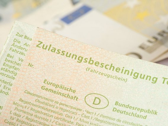 Eine deutsche Fahrzeugzulassungsbescheinigung (Fahrzeugschein) mit grünem Text und dem Länderkennzeichen "D" für Deutschland liegt auf einem Stapel Dokumente.