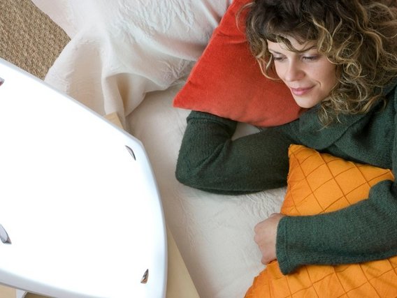 Eine junge Frau mit lockigem Haar liegt auf dem Bett. In ihrer Hand hält sie ein oranges Kissen, während ein rotes Kissen unter ihrem Kopf liegt. Sie blickt auf eine weiße Lampe neben sich.