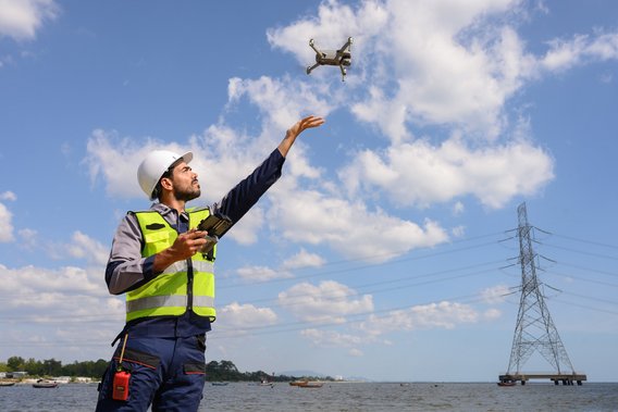 Person in Sicherheitskleidung steuert eine Drohne am Wasser, mit einem Strommast im Hintergrund.