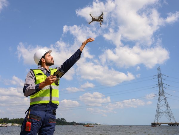 Person in Sicherheitskleidung steuert eine Drohne am Wasser, mit einem Strommast im Hintergrund.