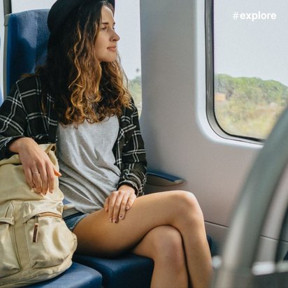 Eine junge Frau mit lockigen Haaren sitzt in einem Zug und schaut aus dem Fenster. Sie trägt ein graues T-Shirt, Shorts und einen schwarzen Hut. Ihre Hand ruht auf ihrer beigen Tasche, die auf dem Sitz neben ihr liegt.