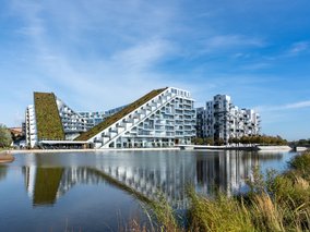 Moderne Gebäude mit begrünten Dächern und schrägen Fassaden spiegeln sich in einem ruhigen Wasserbecken wider.