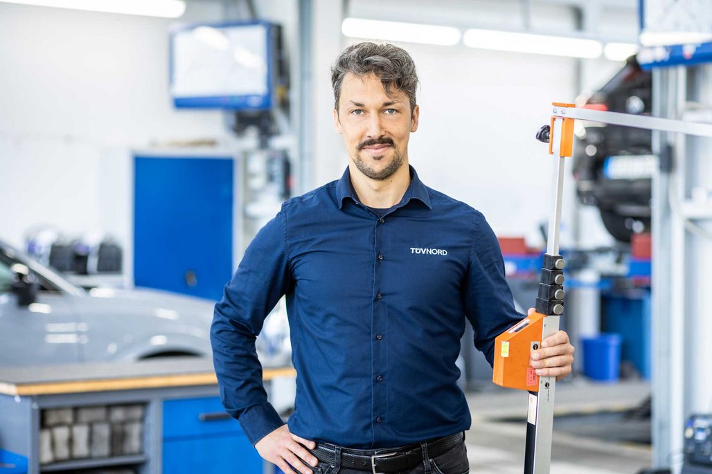 TÜV NORD Mitarbeiter mit Messgerät in einer Werkstatt, im Hintergrund Werkstattausrüstung und ein Auto.