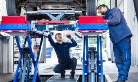 Zwei TÜV NORD Mitarbeiter inspizieren die Unterseite eines Autos auf einer Hebebühne in der Werkstatt.