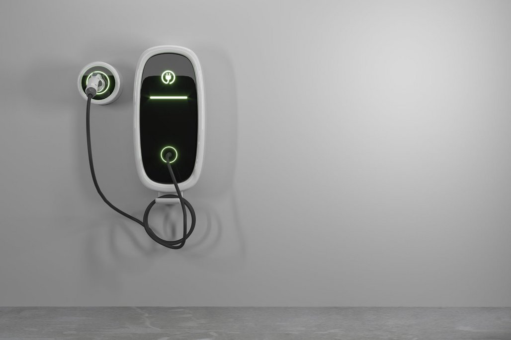 Eine moderne Elektroauto-Ladestation an einer weißen Wand montiert, mit grün leuchtenden Statusanzeigen. Die weiße Wandladestation ist mit einem schwarzen Ladekabel verbunden und zeigt durch die grünen Lichter ihren aktiven Betriebszustand an.