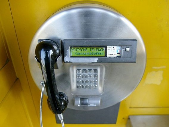 Ein modernes, silbernes Kartentelefon der Deutschen Telekom in einer gelben Telefonzelle. Der Bildschirm zeigt den Schriftzug „Kartentelefon“. Das schwarze Telefon ist mit einem Metallkabel gesichert.