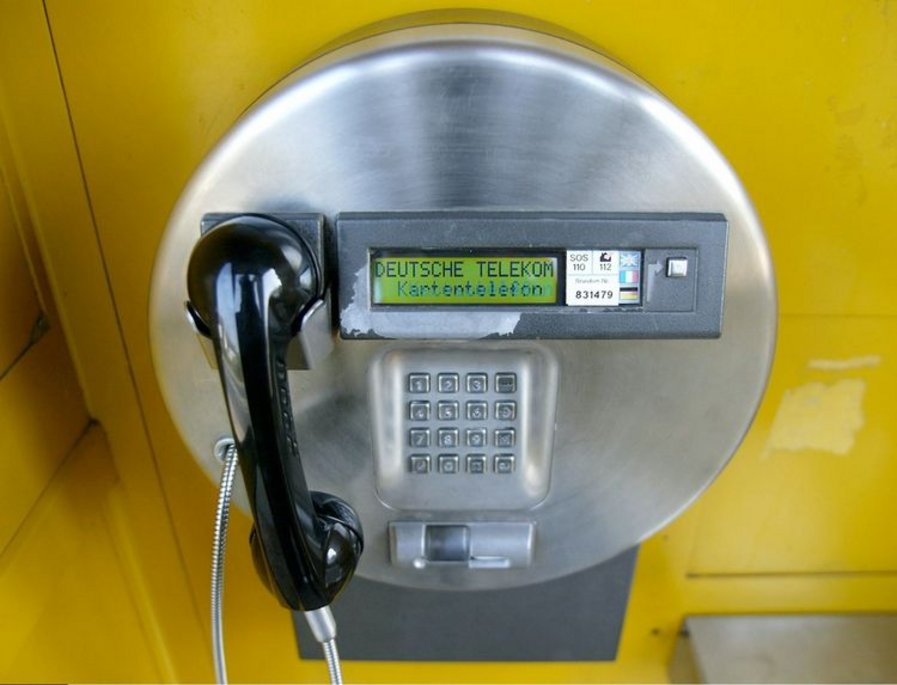 Ein modernes, silbernes Kartentelefon der Deutschen Telekom in einer gelben Telefonzelle. Der Bildschirm zeigt den Schriftzug „Kartentelefon“. Das schwarze Telefon ist mit einem Metallkabel gesichert.