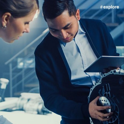 Ein Mann im Anzug und eine Frau im weißen Kittel begutachten gemeinsam einen Motorblock in einer modernen Industriehalle. Die Szene zeigt die Zusammenarbeit zwischen Technik und Management in der Automobilindustrie.