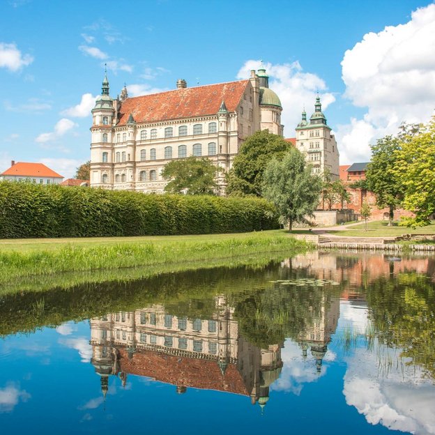 Schloss Güstrow mit Spiegelung im Wasser, umgeben von grüner Landschaft und blauem Himme