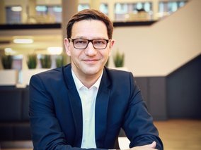 Silvio Konrad mit Brille im blauen Anzug sitzt in einem modernen Büro. Er wirkt professionell und freundlich.