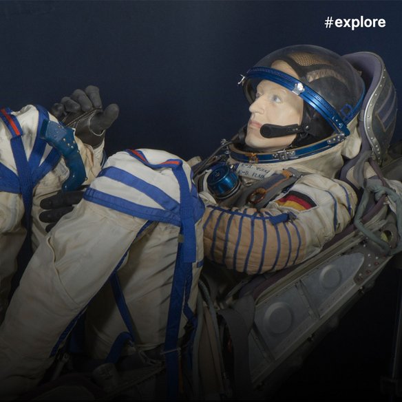 Ein realistisches Plastikmannequin eines Mannes, der eine beige Astronautenuniform mit blauen Linien und einer deutschen Flagge trägt. Er hat ein Mikrofon und schwarze Handschuhe.