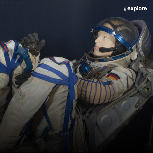 Ein realistisches Plastikmannequin eines Mannes, der eine beige Astronautenuniform mit blauen Linien und einer deutschen Flagge trägt. Er hat ein Mikrofon und schwarze Handschuhe.