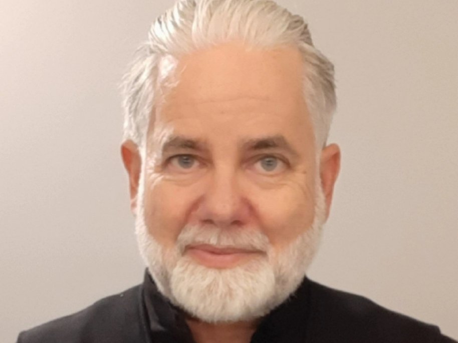 Helge Schmidt mit weißen Haaren und Bart trägt schwarze Kleidung und blickt direkt in die Kamera vor einem neutralen Hintergrund.