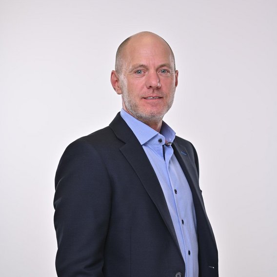 Carsten Becker im dunklen Anzug und hellblauem Hemd steht vor einem weißen Hintergrund in professioneller Pose.